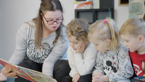 Eine junge Frau mit Trysomie-21 liest Kindern ein Buch vor in einer Kindertagesstätte.  
