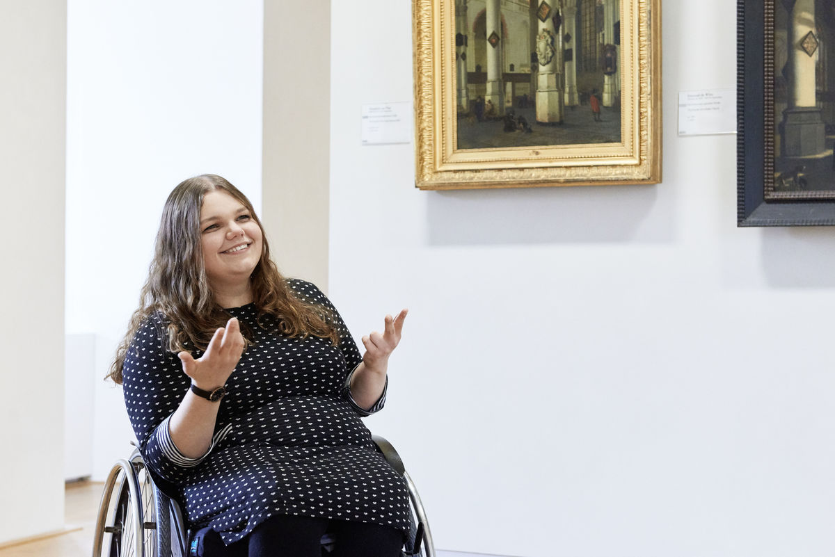 Foto einer lachend gestikulierenden Frau im Rollstuhl in einem Museum, die einen Freiwililgendienst im Kulturbereich leistet